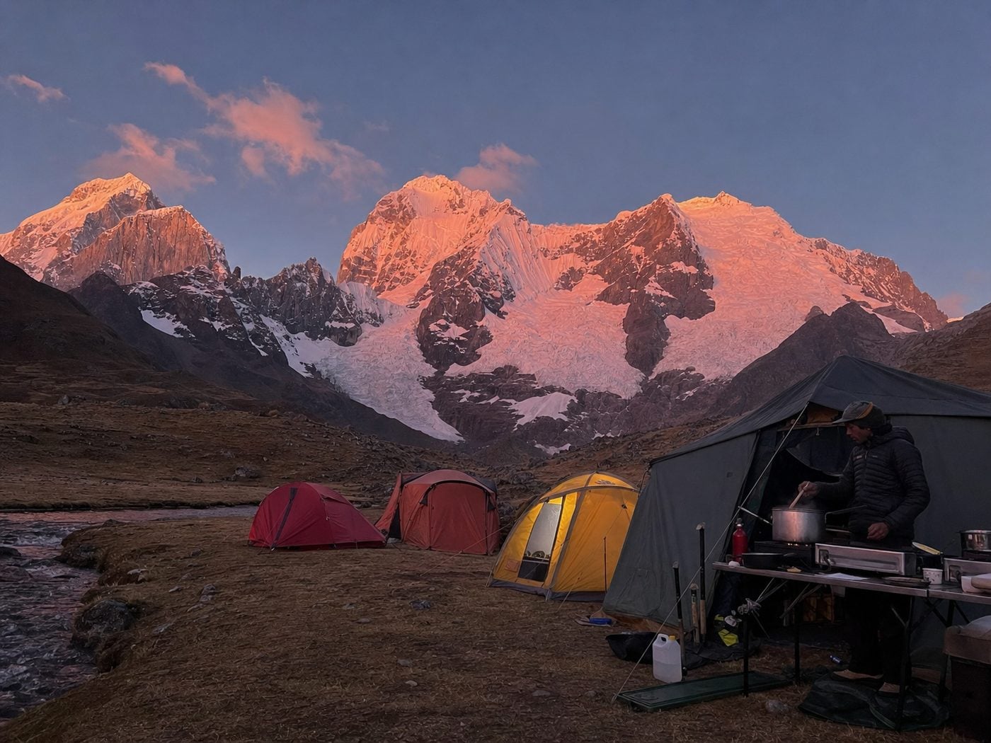 Camp at dusk — tents pitched by a stream, the camp cook working under an awning, alpenglow on the peaks behind.
