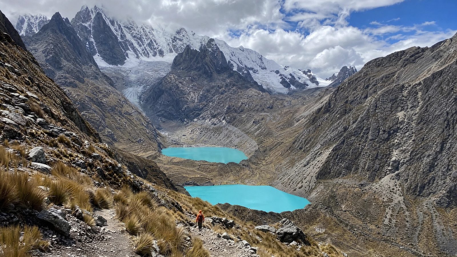 A single trekker on the trail high above two turquoise lagoons, glaciated peaks beyond.