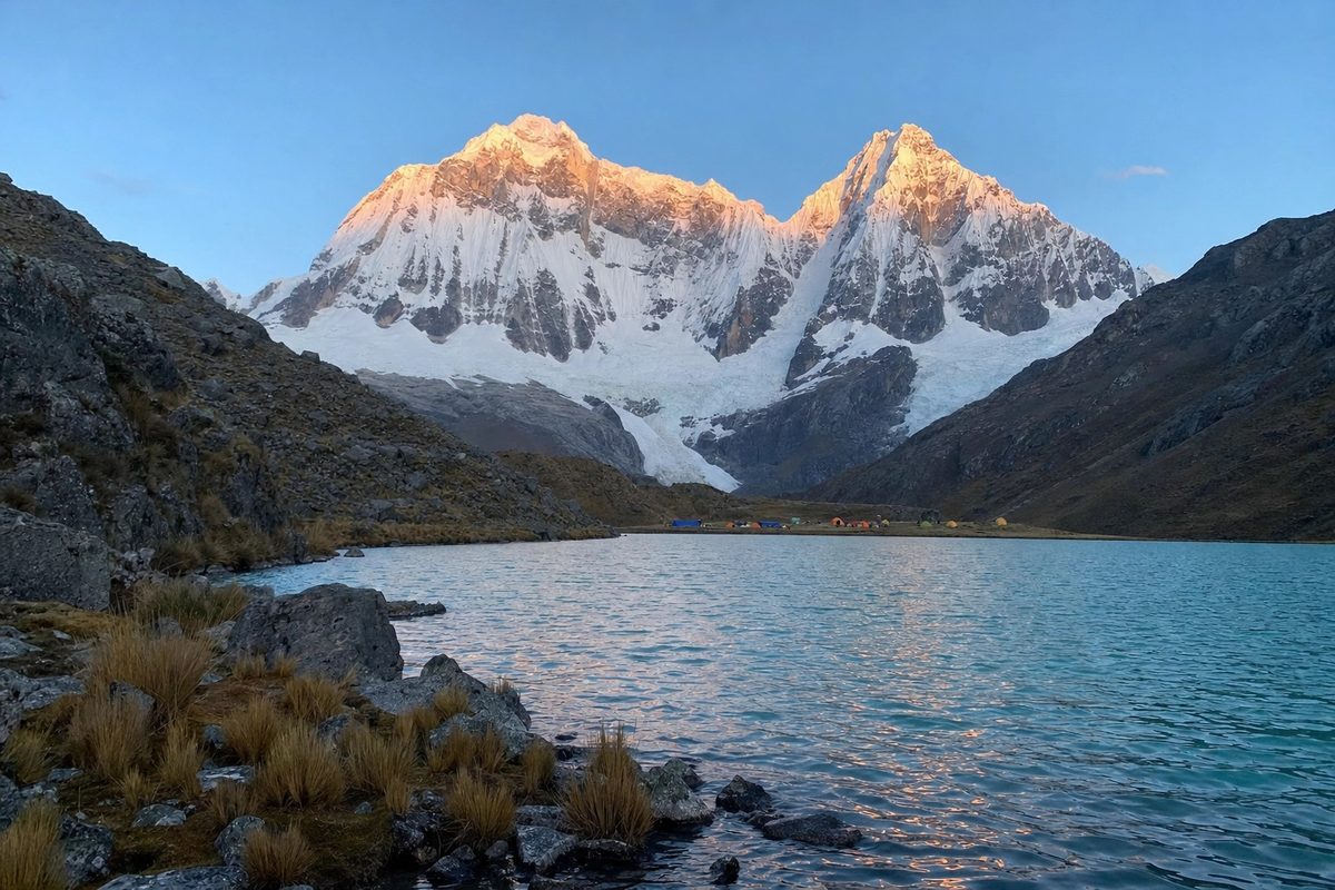 Dawn light striking the peaks above a turquoise lake, camp tents visible on the far shore.