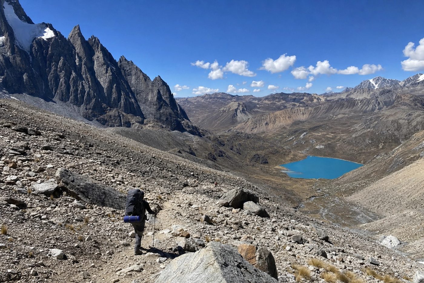 A lone trekker crossing a vast scree slope high above a distant turquoise lake.